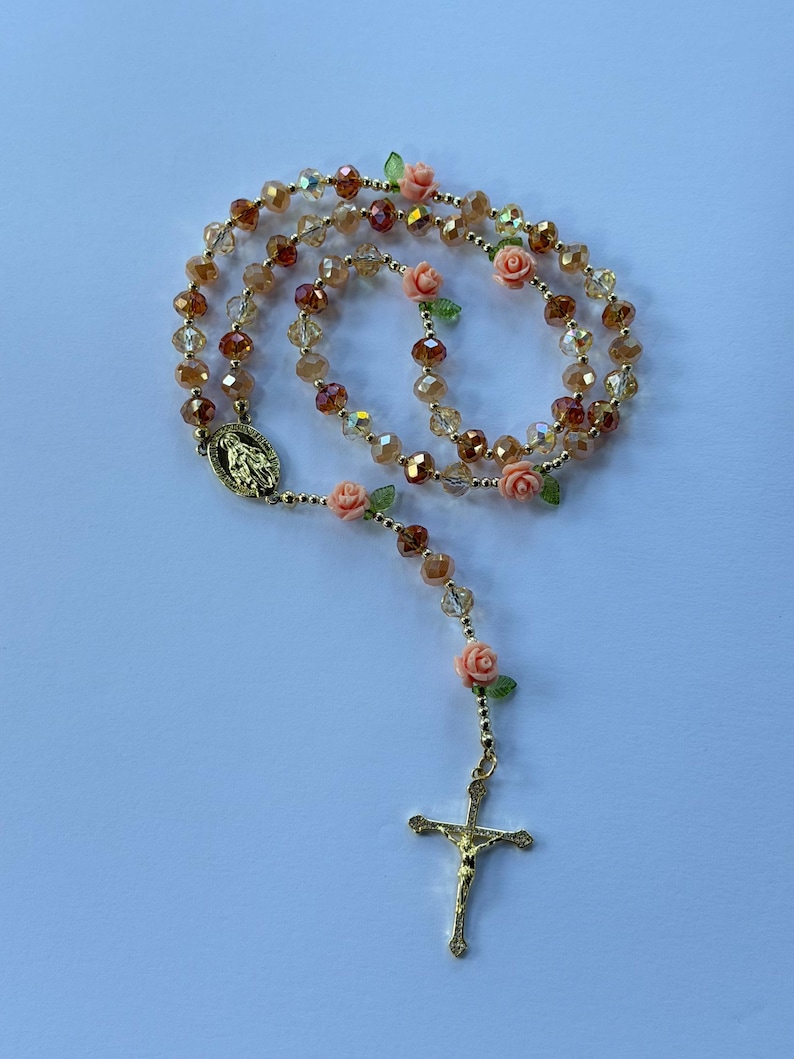 May include: A rosary featuring gold, amber, and brown faceted beads. Small peach-coloured rose accents are placed along the chain. A gold crucifix and a religious medallion are at the end of the chain.