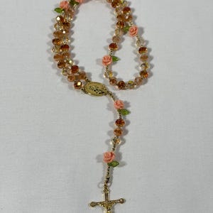 May include: A rosary featuring amber crystal beads and pink rose details. The rosary has a gold-coloured crucifix and a gold-coloured centrepiece with an oval design. The beads are strung together with gold-coloured wire.