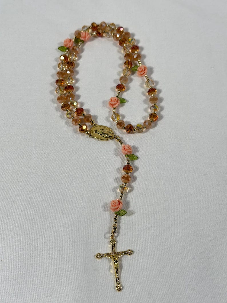 May include: A rosary featuring amber crystal beads and pink rose details. The rosary has a gold-coloured crucifix and a gold-coloured centrepiece with an oval design. The beads are strung together with gold-coloured wire.