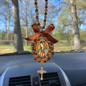 May include: A brown and gold beaded car charm with a picture of the Virgin Mary and a cross. The charm is hanging from the rearview mirror of a car.