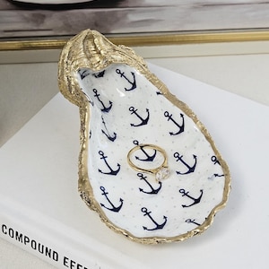 May include: A gold-edged oyster shell ring dish with a white interior patterned with navy blue anchors. A gold ring with a clear stone rests inside the dish. The dish sits on a white book with the title "Compound Effect".