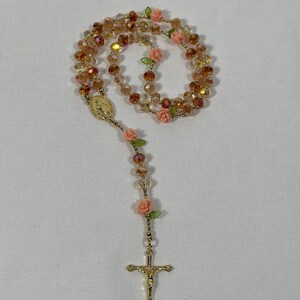 May include: A rosary featuring amber-coloured beads and gold-tone details. It includes small pink rose accents and a gold crucifix. A gold medallion is also present.