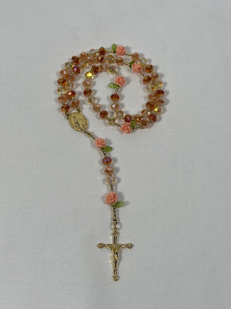 May include: A rosary featuring amber-coloured beads and gold-tone details. It includes small pink rose accents and a gold crucifix. A gold medallion is also present.
