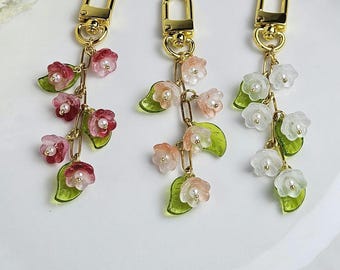 May include: Three floral keychains with gold-colored hardware. Each keychain features a chain of small, translucent flowers in shades of pink, peach, and white, with green leaves and faux pearls. The keychains are arranged on a white surface.