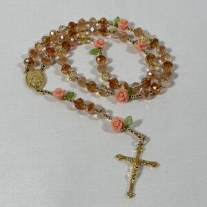May include: A rosary featuring amber-coloured faceted beads and small pink rose accents. It has a gold-coloured crucifix and a medallion with an embossed image. The beads are strung on a gold-coloured chain.