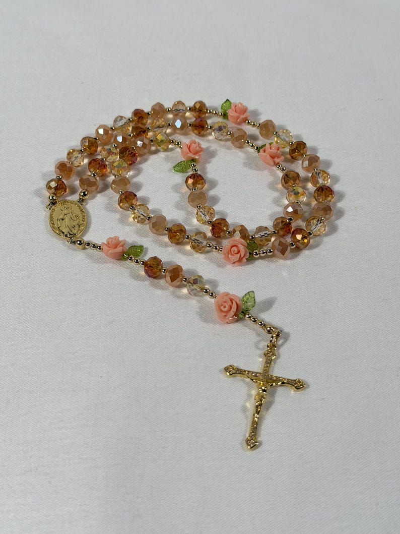 May include: A rosary featuring amber-coloured faceted beads and small pink rose accents. It has a gold-coloured crucifix and a medallion with an embossed image. The beads are strung on a gold-coloured chain.