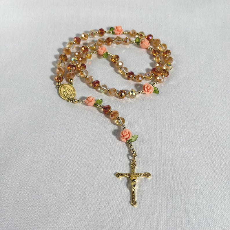 May include: A rosary featuring amber and clear beads, a gold-tone crucifix, and peach-colored rose accents. The rosary includes a gold-tone medallion. The beads are strung together to form a necklace.
