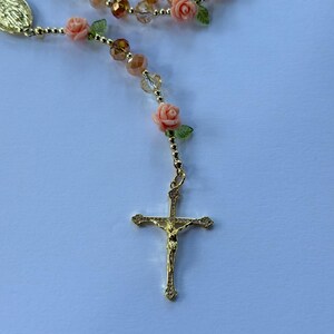 May include: A gold rosary with a crucifix pendant. The rosary features peach rose beads, green leaf accents, and clear and amber crystal beads. The rosary is designed for prayer and religious devotion.