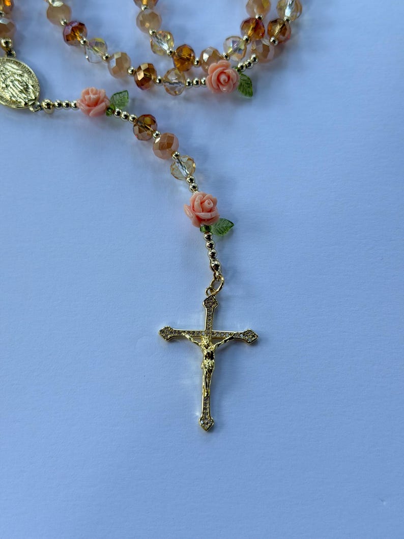 May include: A gold rosary with a crucifix pendant. The rosary features peach rose beads, green leaf accents, and clear and amber crystal beads. The rosary is designed for prayer and religious devotion.