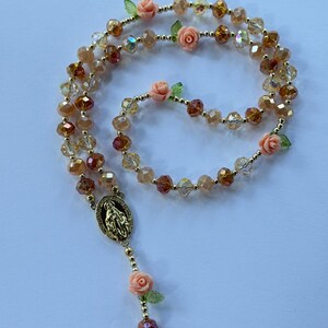 May include: A rosary featuring faceted amber and clear beads, decorated with small peach-coloured rose charms and green leaves. A gold-toned pendant with a religious figure is at the centre. The beads are strung on a gold-coloured chain.