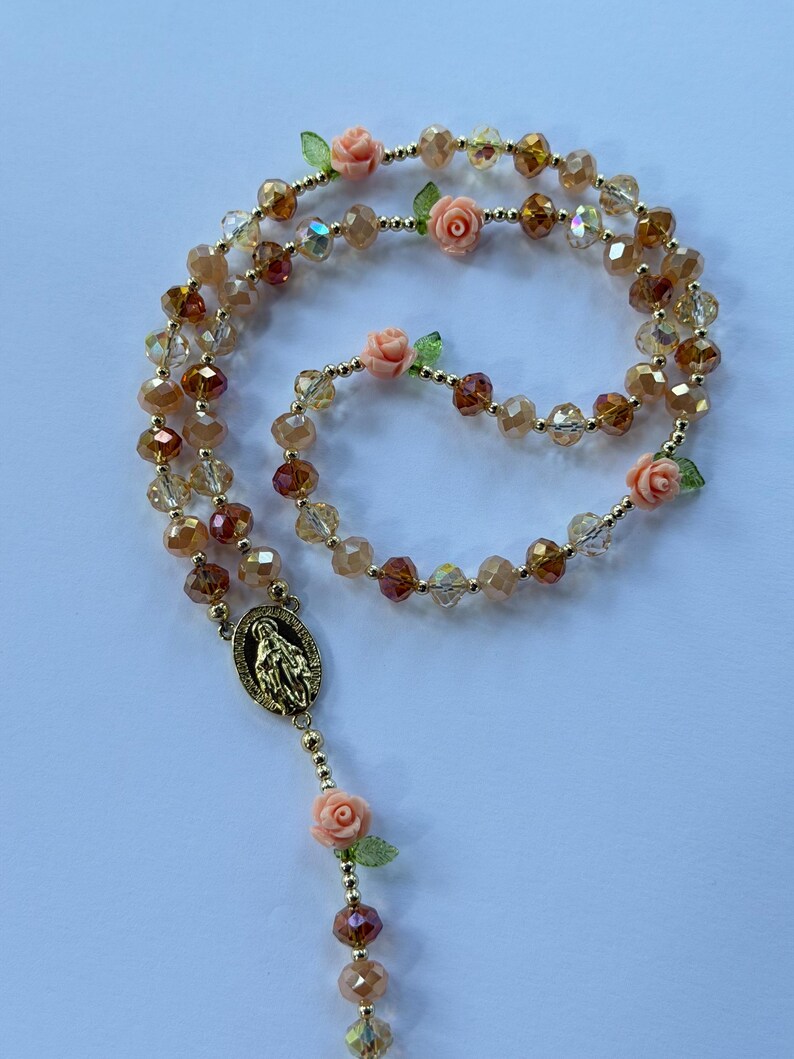 May include: A rosary featuring faceted amber and clear beads, decorated with small peach-coloured rose charms and green leaves. A gold-toned pendant with a religious figure is at the centre. The beads are strung on a gold-coloured chain.