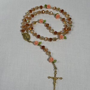 May include: A rosary featuring amber-toned faceted beads and small pink rose embellishments. It includes a gold-coloured crucifix and a religious medallion. The beads are strung together with gold-coloured wire.