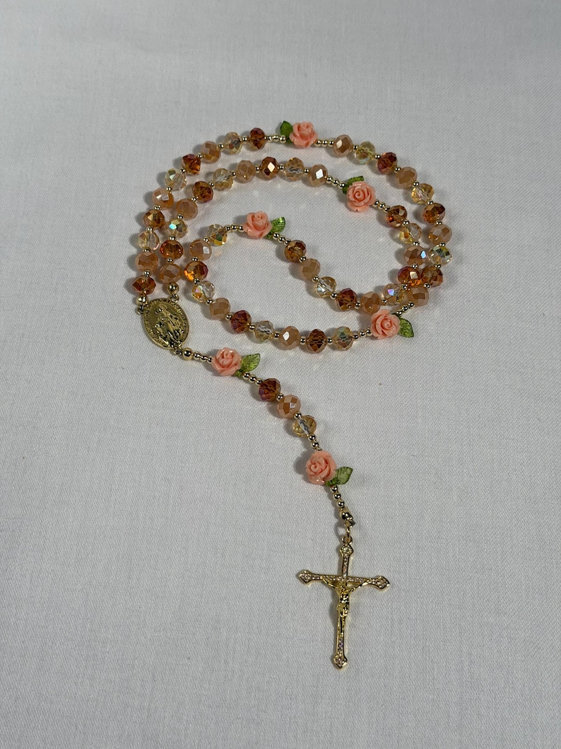 May include: A rosary featuring amber-toned faceted beads and small pink rose embellishments. It includes a gold-coloured crucifix and a religious medallion. The beads are strung together with gold-coloured wire.
