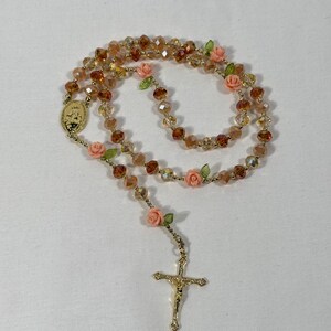 May include: A rosary featuring amber-coloured crystal beads and gold-tone metal detailing. The rosary includes small pink rose accents and a gold crucifix pendant. The beads are strung on a gold-coloured chain.