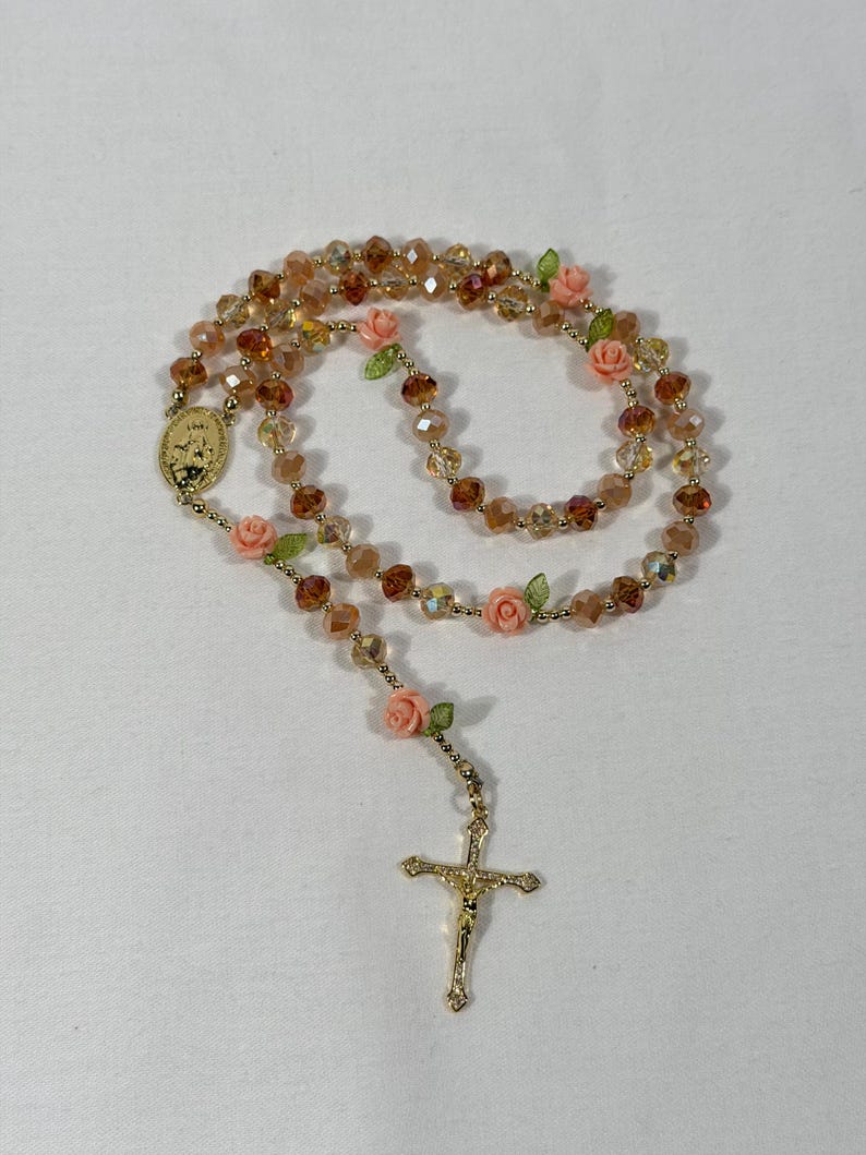 May include: A rosary featuring amber-coloured crystal beads and gold-tone metal detailing. The rosary includes small pink rose accents and a gold crucifix pendant. The beads are strung on a gold-coloured chain.
