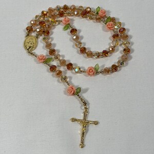 May include: A rosary featuring amber-toned faceted beads, with small pink rose and green leaf details. It has a gold-coloured crucifix and a medallion with a religious figure.