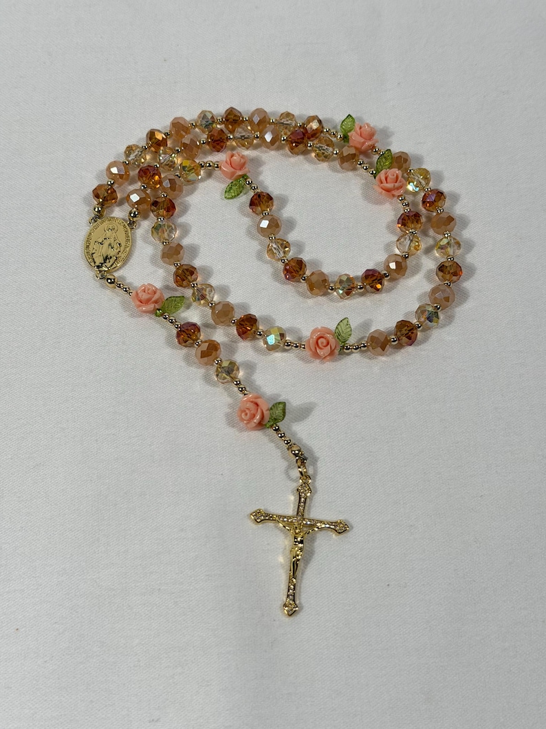 May include: A rosary featuring amber-toned faceted beads, with small pink rose and green leaf details. It has a gold-coloured crucifix and a medallion with a religious figure.