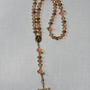 May include: A rosary featuring amber-toned faceted beads and small pink rose embellishments. It includes a gold-coloured crucifix and a religious medallion. The beads are strung on a gold-coloured chain.