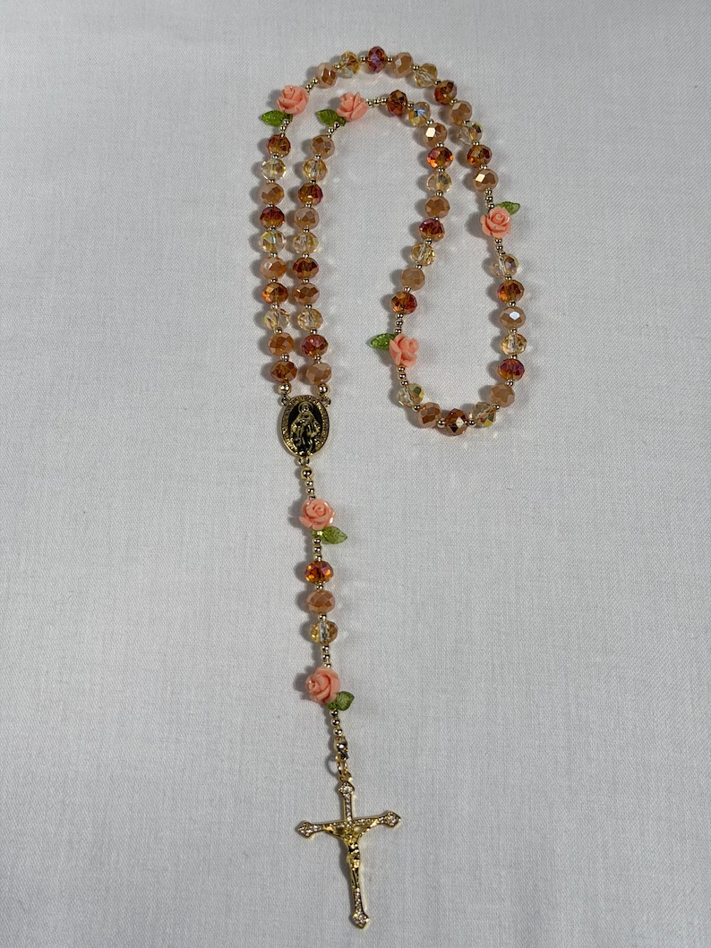 May include: A rosary featuring amber-toned faceted beads and small pink rose embellishments. It includes a gold-coloured crucifix and a religious medallion. The beads are strung on a gold-coloured chain.