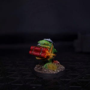 May include: A miniature figurine of a green creature with a gray hat, holding a bundle of red dynamite. The figure is painted with orange and yellow accents and stands on a small, textured base with grass.