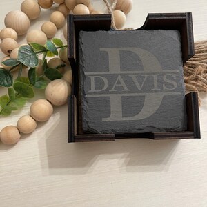 May include: A black slate coaster with the letter "D" and the name "DAVIS" engraved in white. The coaster is in a wooden holder.