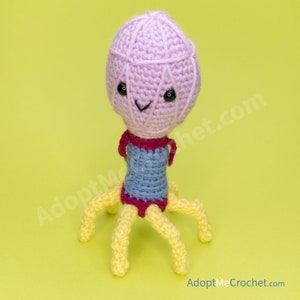 May include: A crocheted bacteriophage toy with a pink head, blue body, and yellow legs. The toy has black eyes and a smiling mouth.