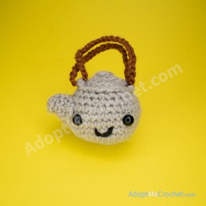 May include: A crocheted teapot with a brown handle and a smiling face. The teapot is gray and has black eyes.