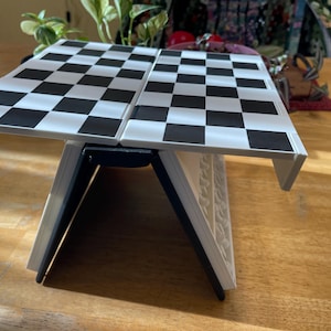 Puede incluir: Un tablero de ajedrez plegable con un patrón de cuadros blancos y negros. El tablero está sostenido por un marco blanco y negro, creando una configuración de juego de mesa. El tablero está abierto y listo para jugar.