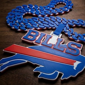 May include: A large, flat, three-dimensional pendant featuring the word Bills in blue letters with red outlines, positioned above a blue and red buffalo logo. The pendant is attached to a thick, chunky chain made of interlocking blue and white links.