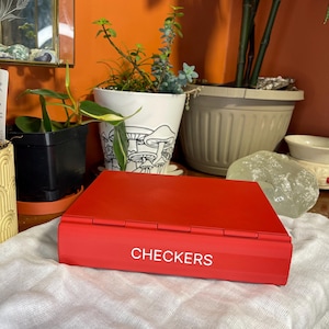 Puede incluir: Una caja de juego de damas de color rojo brillante con la palabra "CHECKERS" en letras blancas. La caja rectangular está sobre una tela blanca. El fondo incluye plantas en macetas y una pared naranja.