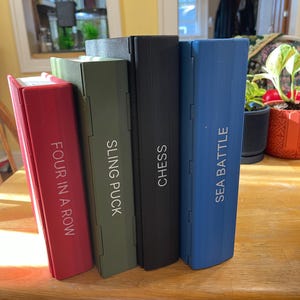 Puede incluir: Cuatro cajas de juegos coloridas se muestran sobre una mesa de madera. Las cajas son rojas, verde oliva, negras y azules. Cada caja tiene texto blanco con los nombres de los juegos: "FOUR IN A ROW", "SLING PUCK", "CHESS" y "SEA BATTLE".