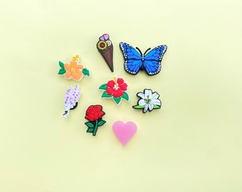May include: Assortment of colorful flower and butterfly shoe charms. The charms are made of rubber and feature a variety of designs, including a hibiscus, a rose, a lily, a butterfly, and a bouquet of flowers.