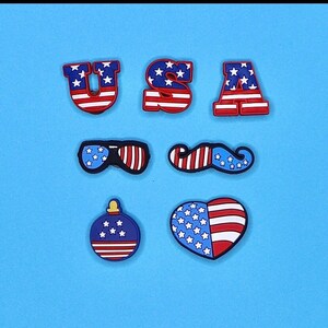 May include: A set of seven red, white, and blue shoe charms featuring the letters "USA", sunglasses, a mustache, a heart, and an ornament. The charms are decorated with stars and stripes.