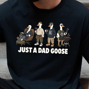 May include: Black t-shirt with the text "JUST A DAD GOOSE" in white. The graphic features four cartoon geese dressed as a dad in different scenarios: relaxing, working, drinking coffee, and grilling.