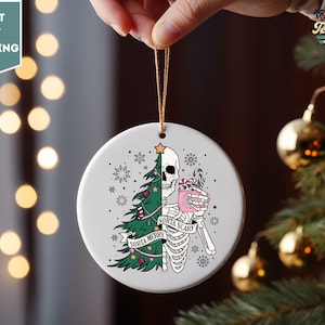May include: White ceramic Christmas ornament with a skeleton holding a pink drink and a Christmas tree growing out of its ribcage. The text on the ornament reads "Sorta Merry Sorta Scary".