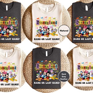 Peut inclure: Plusieurs t-shirts avec un motif Disneyland coloré, le texte "Disneyland 2026" et un espace pour un nom. Disponibles en gris foncé, naturel et gris chiné foncé. Les t-shirts sont présentés sur une surface tressée.