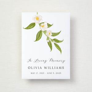 May include: A white card with a floral illustration and text. The illustration features white flowers and green leaves. The text reads "In Loving Memory" followed by a name and dates: May 17, 1923 - June 5, 2025.