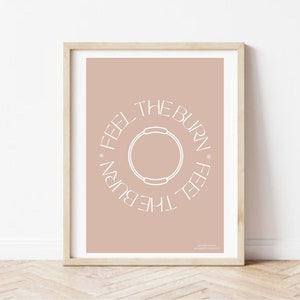 Puede incluir: Una impresión enmarcada con un fondo rosa claro y un círculo blanco con el texto "Feel the Burn" repetido alrededor del círculo.