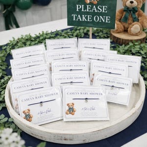 May include: A baby shower display featuring a sign with a teddy bear illustration and the text "PLEASE TAKE ONE." A wooden tray holds several small cards with bracelets, each card featuring a teddy bear and the words "CASEYS BABY SHOWER."