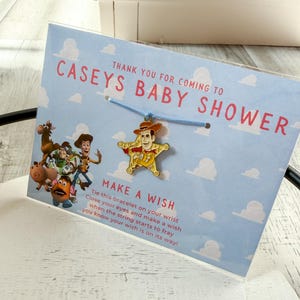 Op de afbeelding: Een babyshower gunst met een Woody bedelarmband. De bedel heeft de vorm van een ster met een Woody figuur. De kaart leest "CASEYS BABY SHOWER" en "MAKE A WISH". De achtergrond is lichtblauw met witte wolken.