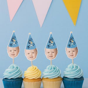 May include: Four cupcakes with blue frosting and blue cupcake liners, and one with yellow frosting and a yellow cupcake liner. Each cupcake has a topper with a baby's face wearing a blue party hat. A pink, white, and yellow banner is in the background.