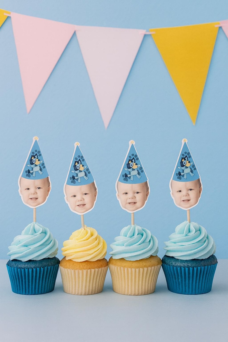 May include: Four cupcakes with blue frosting and blue cupcake liners, and one with yellow frosting and a yellow cupcake liner. Each cupcake has a topper with a baby's face wearing a blue party hat. A pink, white, and yellow banner is in the background.