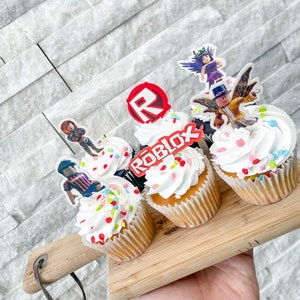 Puede incluir: Cuatro cupcakes decorados con glaseado blanco y chispas de colores. Cada cupcake tiene una decoraci&oacute;n diferente de un personaje de Roblox. Los cupcakes est&aacute;n sobre una tabla de cortar de madera.
