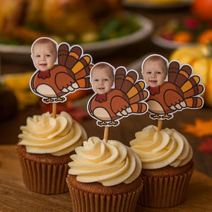 May include: Three cupcakes with light yellow frosting and turkey-themed toppers featuring a baby's face. The cupcakes sit on a wooden board, with a roasted turkey and other Thanksgiving foods in the background.