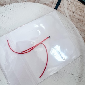 Puede incluir: Una bolsa de pl&aacute;stico transparente con un hilo rojo en su interior. La bolsa est&aacute; sobre una superficie de madera blanca.
