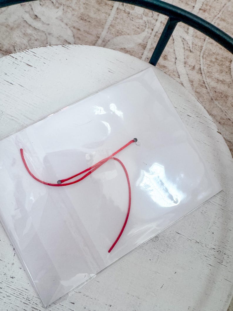 Puede incluir: Una bolsa de pl&aacute;stico transparente con un hilo rojo en su interior. La bolsa est&aacute; sobre una superficie de madera blanca.