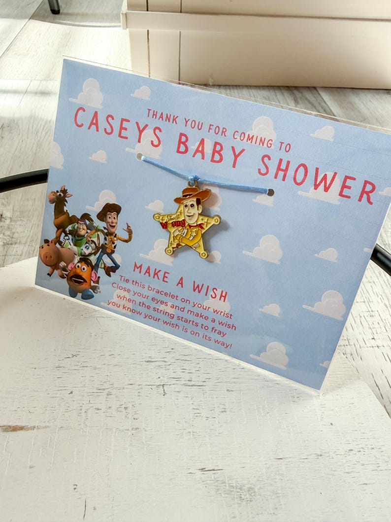 Op de afbeelding: Een babyshower gunst met een Woody bedelarmband. De bedel heeft de vorm van een ster met een Woody figuur. De kaart leest "CASEYS BABY SHOWER" en "MAKE A WISH". De achtergrond is lichtblauw met witte wolken.