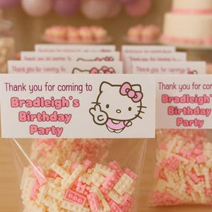 以下が含まれることがあります： ピンクと白のキャンディーレンガが入った透明なセロハン袋。 「Bradleigh's Birthday Partyへようこそ」と書かれたサインには、ハローキティのグラフィックが描かれています。 背景には風船とケーキがあります。