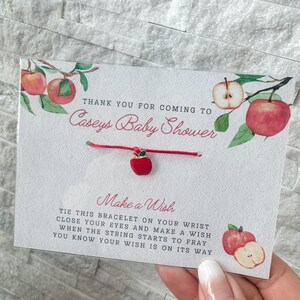 Puede incluir: Pulsera roja con un dije de manzana roja y verde y una tarjeta blanca que dice "Gracias por venir a la fiesta de bienvenida de Caseys Make a Wish Ata esta pulsera en tu mu&ntilde;eca cierra los ojos y pide un deseo cuando la cuerda empiece a deshilacharse sabr&aacute;s que tu deseo est&aacute; en camino"