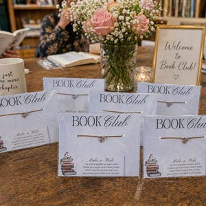 Può includere: Un'ambientazione da tavolo per un book club, con biglietti "Book Club" con braccialetti, un cartello incorniciato con la scritta "Benvenuti al Book Club!", una tazza con testo, fiori e libri.